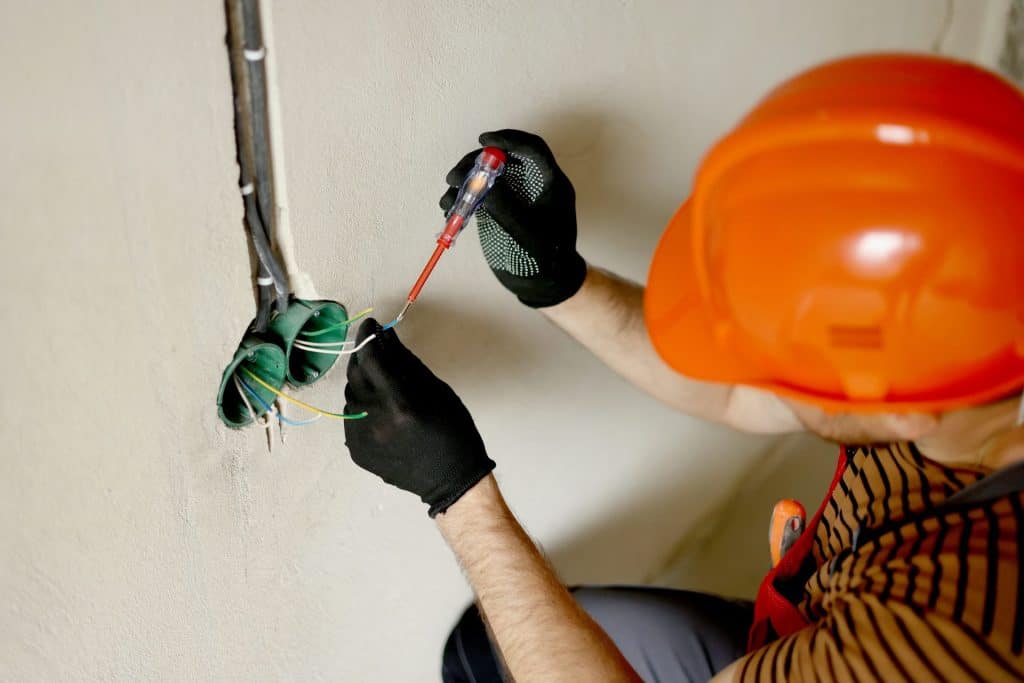 Un électricien portant un casque de sécurité orange et des gants utilise un tournevis pour travailler sur le câblage électrique d'un mur lors d'une rénovation électrique.