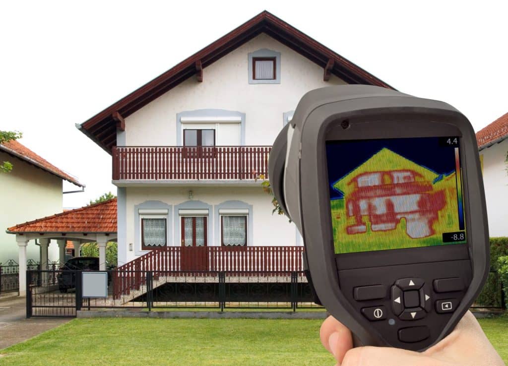 Une personne utilise une caméra thermique pour capturer une carte thermique d'une maison à deux étages, révélant les ponts thermiques et les variations de température sur la façade du bâtiment.
