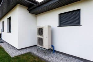 Une pompe à chaleur économique extérieure est installée à côté d'un bâtiment blanc avec une base en gravier et un volet de fenêtre fermé au-dessus.