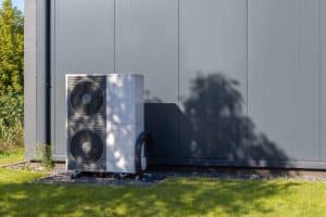 Une pompe à chaleur extérieure blanche avec deux ventilateurs noirs est installée à côté d'un bâtiment moderne gris sur une pelouse, soulignant l'importance de savoir entretenir une pompe à chaleur pour une efficacité durable.