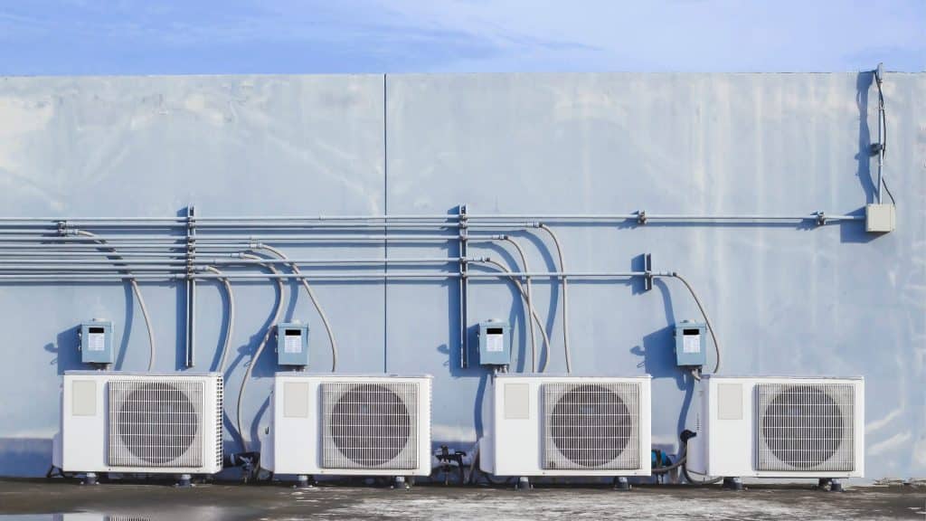 Quatre condenseurs d'air conditionné extérieurs sont installés en rangée contre un mur bleu clair, avec des tuyaux de canalisation électrique qui courent au-dessus d'eux - une inspiration idéale si vous envisagez d'acheter une pompe à chaleur pour votre espace.