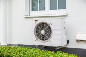 Une unité de climatisation extérieure, idéale pour ceux qui cherchent à acheter une pompe à chaleur, est montée sur le mur extérieur d'un bâtiment sous une fenêtre, avec des arbustes verts au premier plan.